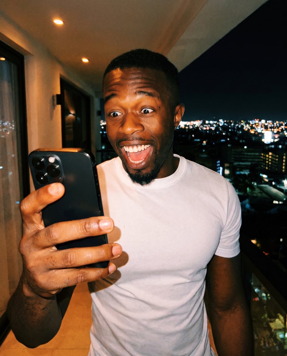 A man on a balcony at night reacting with delight to something on his phone